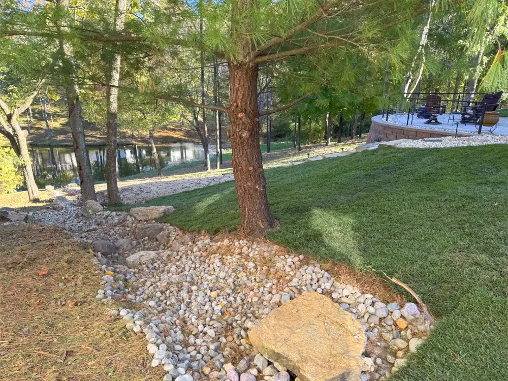 French Drain with rocks surrounding in a landscaped backyard with green grass and a patio area with chairs near a pond.