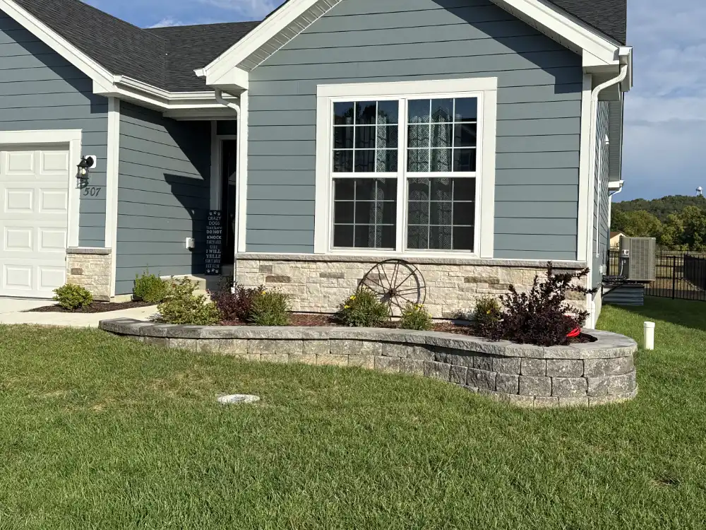 Front yard flower bed with stone retaining wall and decorative wagon wheel in front of a blue house with white window trim.