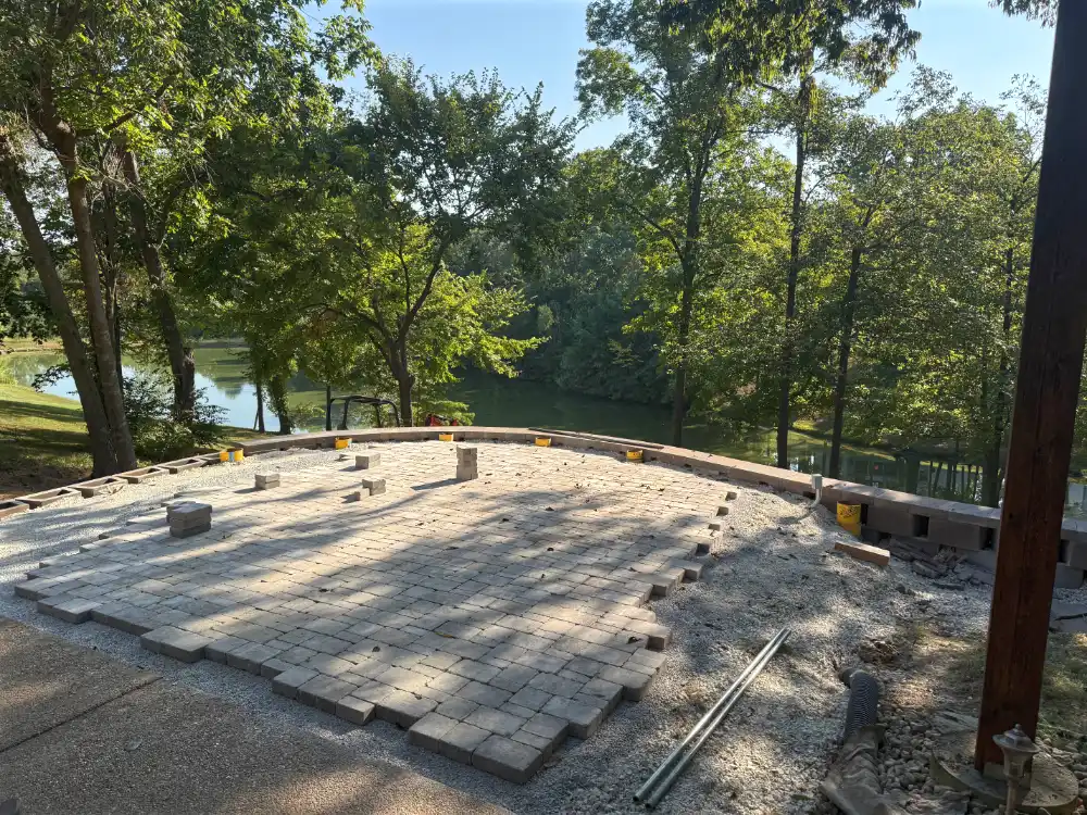 Partially completed stone paver patio with gravel base surrounded by trees near a lake.