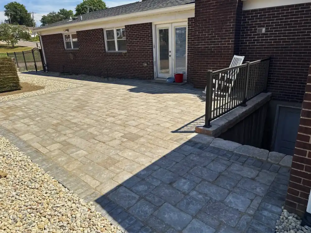 Stone paver patio with light and dark square tiles adjacent to a brick house with white-framed glass doors and a fenced stairway.
