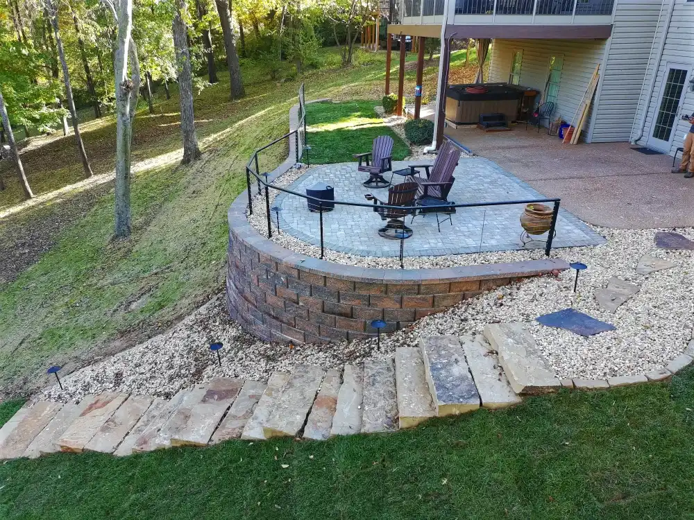 Stone patio with Adirondack chairs and a fire pit, surrounded by a curved retaining wall and a pathway of flat stones through white gravel in a backyard.