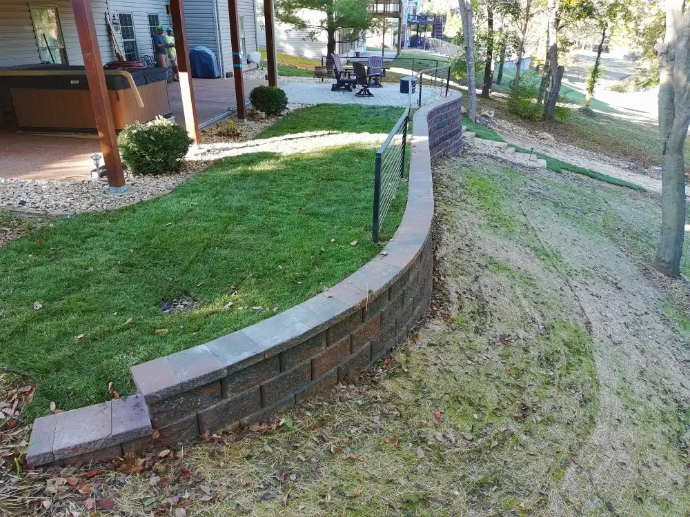 Curved retaining wall separating a grassy patio area with outdoor chairs from a sloped sandy yard with trees.