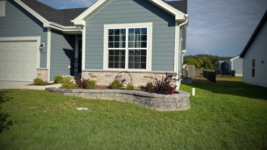 House featuring a stone raised garden bed with various small shrubs and a decorative metal wheel in front.