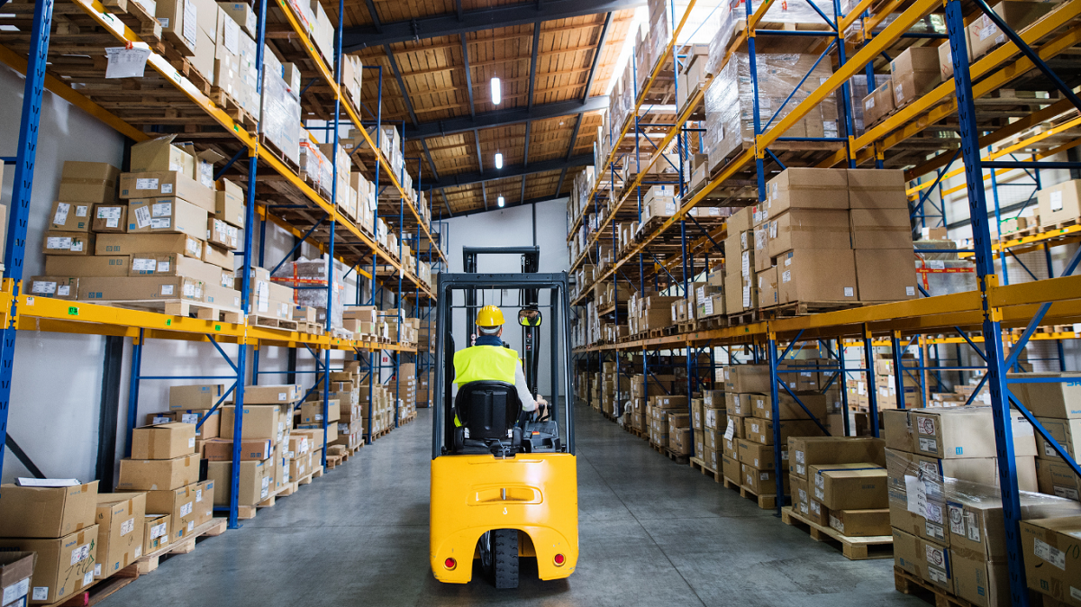 Warehouse worker in a hard hat and hi-vis vest driving a yellow forklift down an aisle between tall pallet racks stocked with boxes in a distribution warehouse.