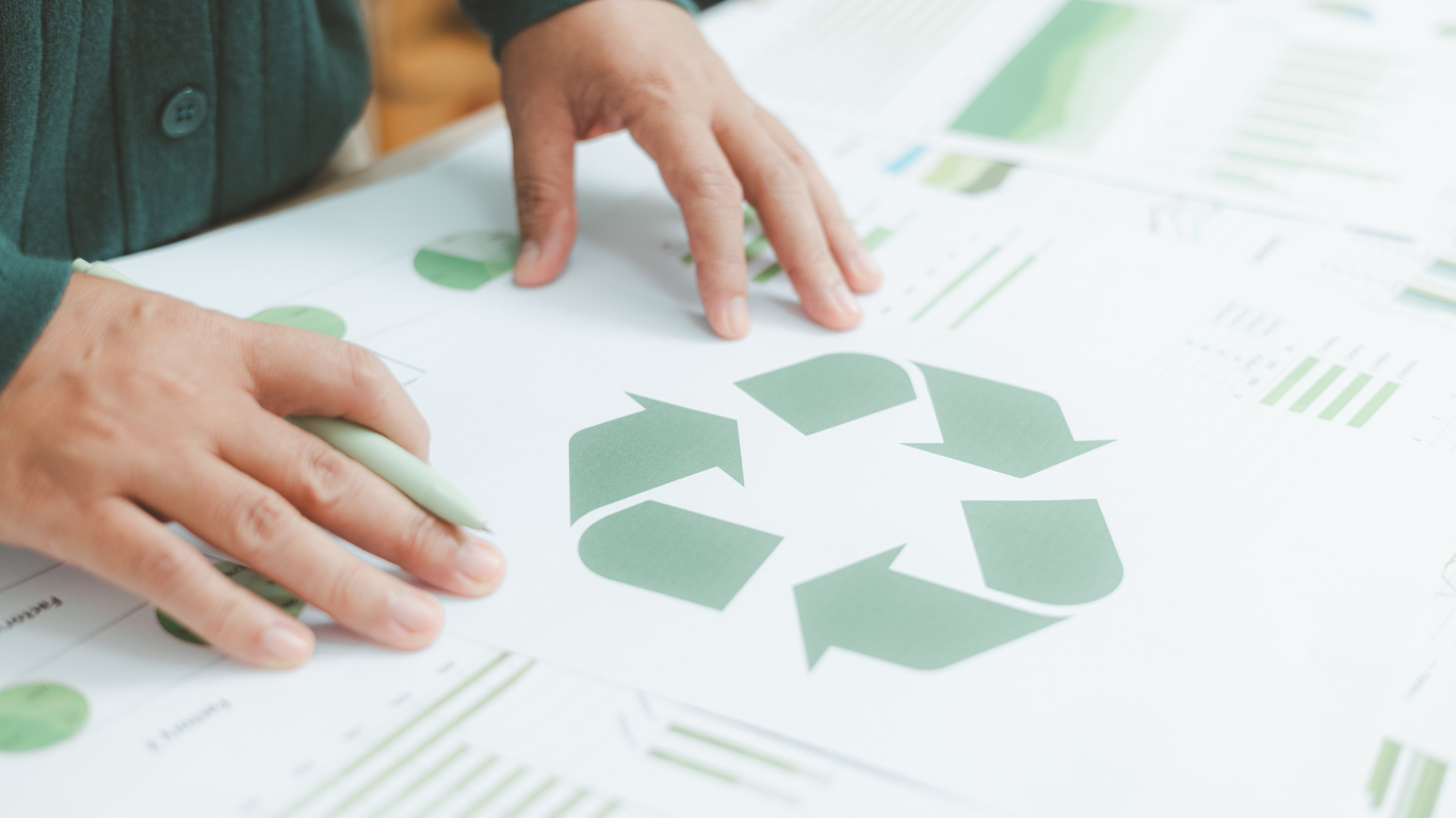 Hands arranging green paper shapes on a table covered with diagrams and labels, with one person placing a cut-out piece over a cluster of recycled-looking paper scraps.