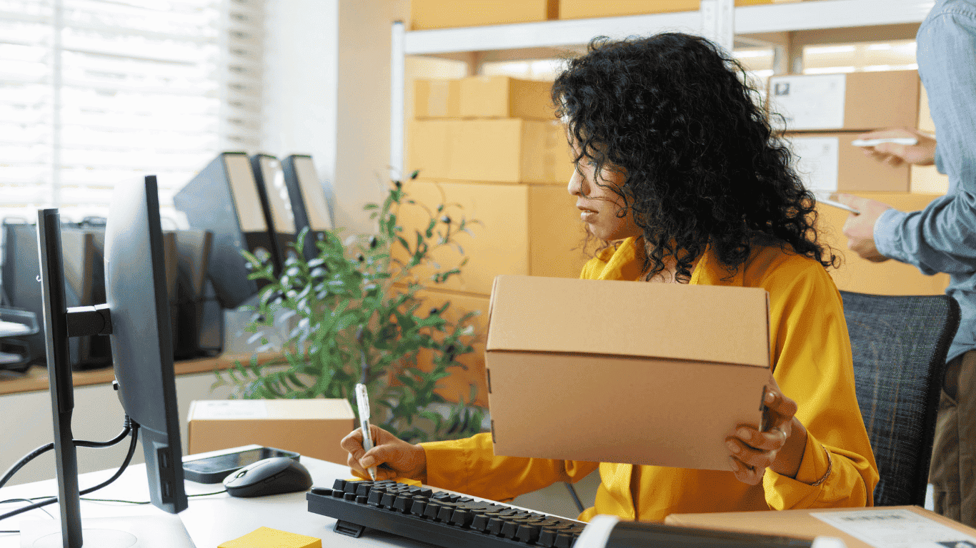 In a bright office, a person in a yellow shirt holds a stack of cardboard boxes while preparing shipments at a desk with a keyboard and desk phone, with more boxes and coworkers in the background.