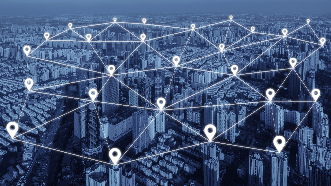 A city skyline viewed from above with glowing network lines and pin icons linking buildings, forming a digital connectivity overlay across the urban landscape.