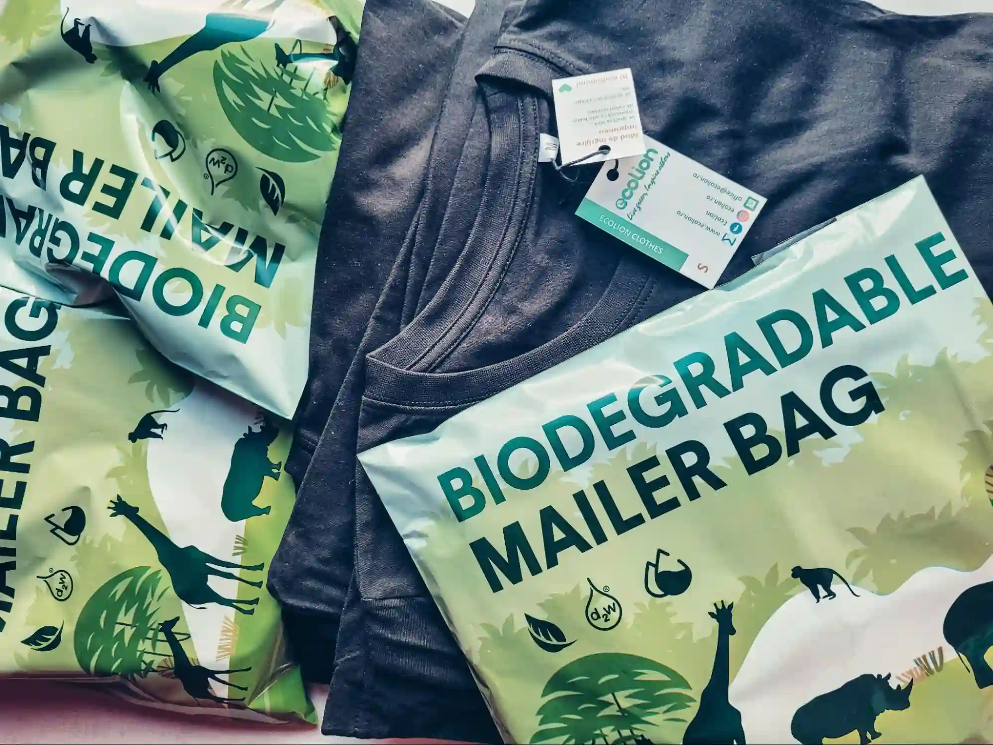 An eco-friendly shipping package containing a dark gray t-shirt with retail tags, surrounded by custom-printed green biodegradable poly mailers featuring a wildlife safari illustration.