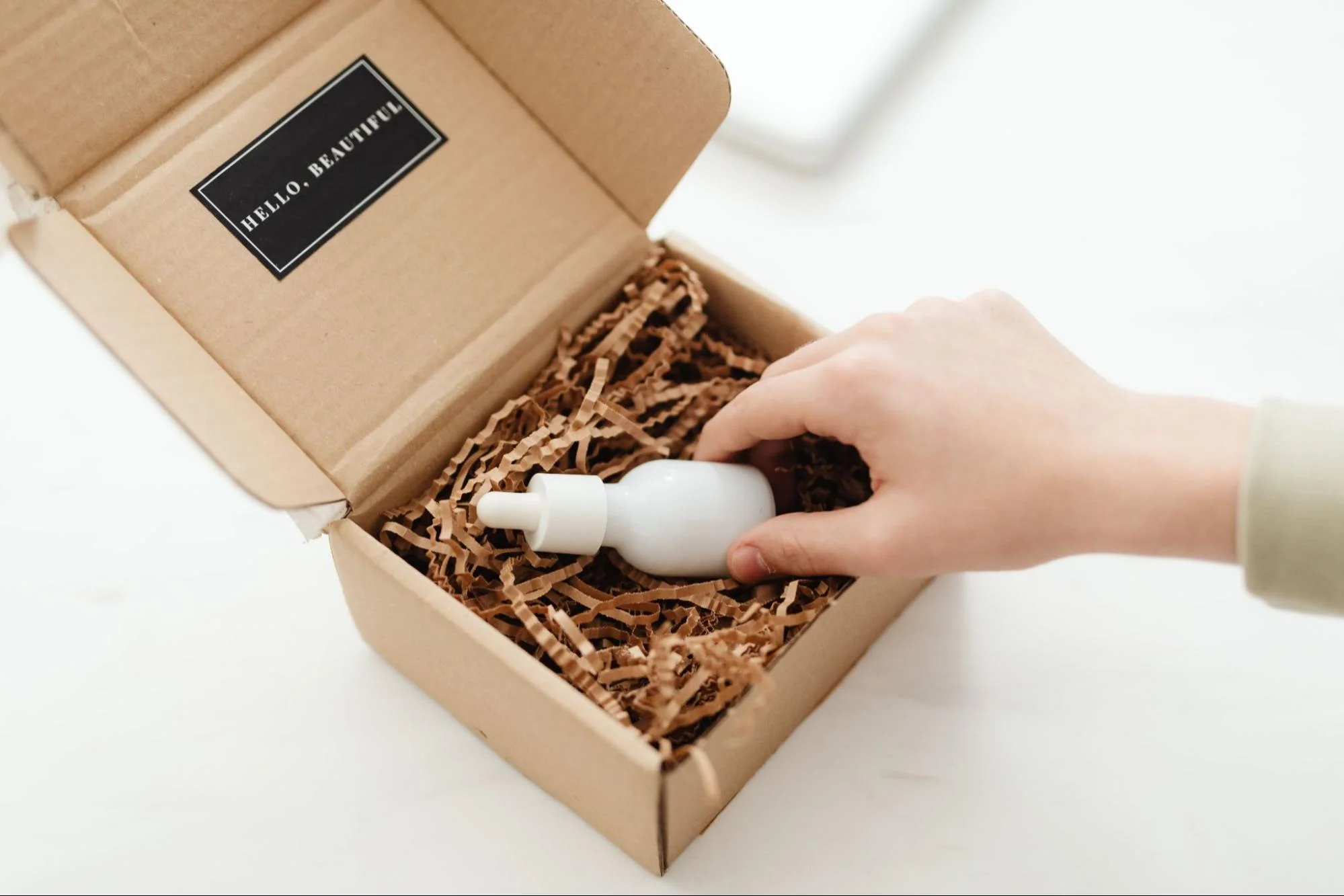 A hand is placing a white glass dropper bottle into a small cardboard box filled with brown crinkle paper filler.