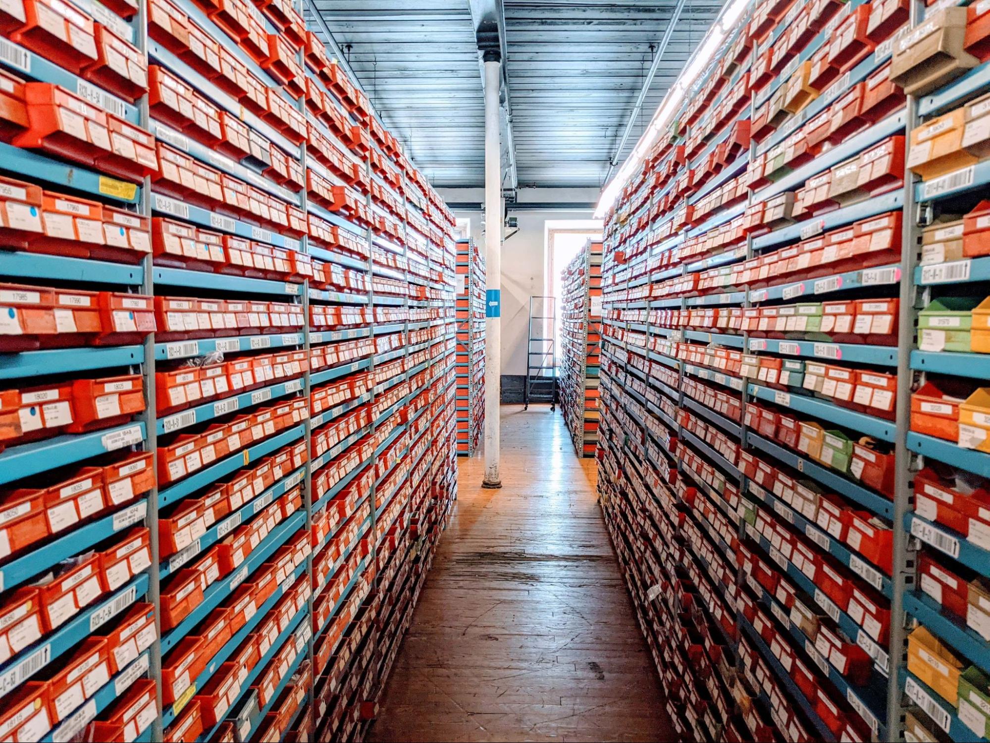 An aisle in a warehouse lined with tall metal shelves containing rows of orange and red storage boxes on a wooden floor.