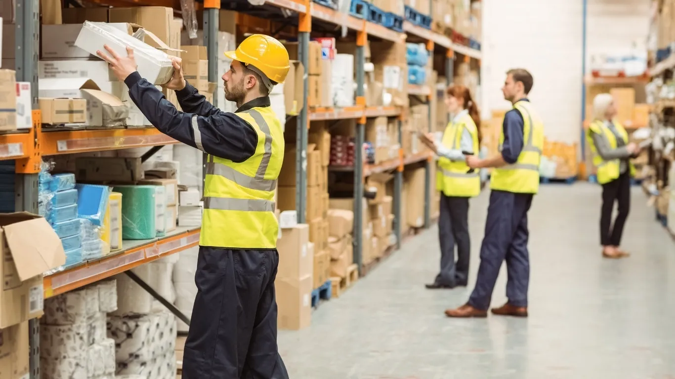 Warehouse workers in yellow safety vests and hard hats move boxes along tall shelves, with one worker handing a package to a coworker in a busy aisle.
