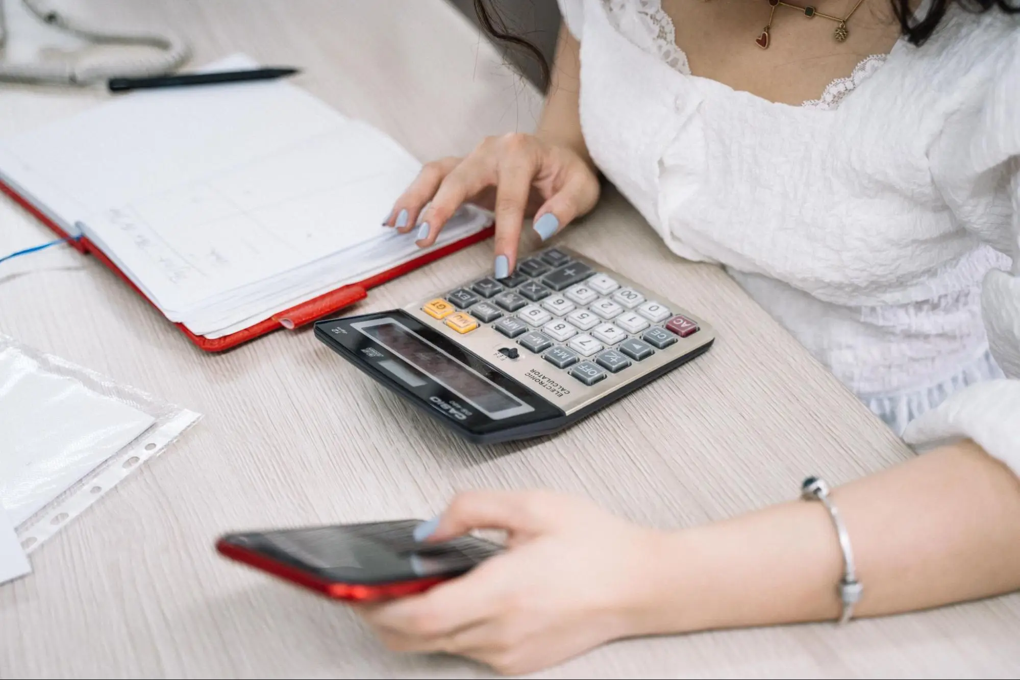 A person in a white blouse using a desktop calculator with one hand and holding a red smartphone with the other, next to an open notebook on a light wood desk.