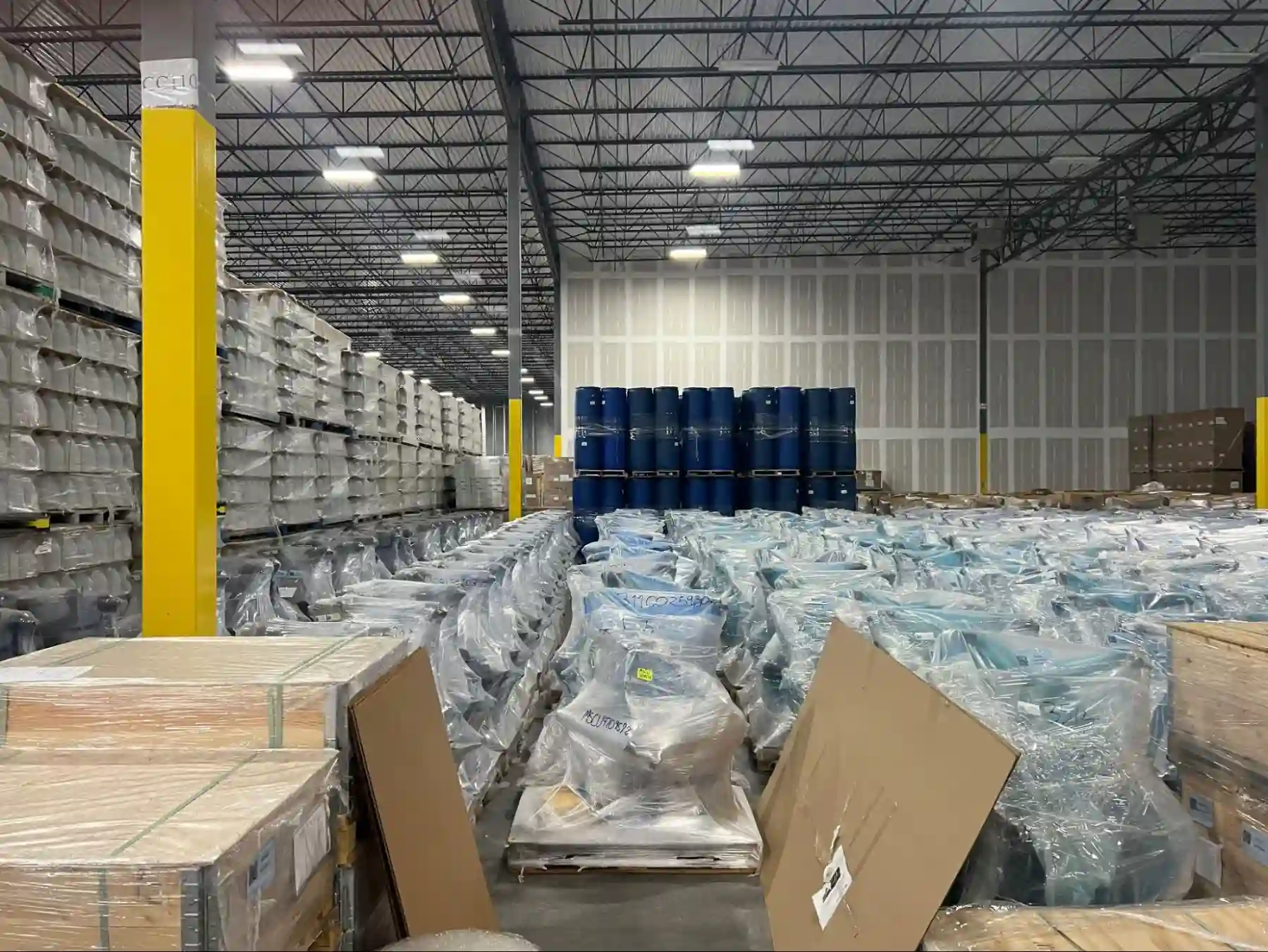 A large warehouse interior with tall racks of shrink-wrapped white containers, rows of plastic-wrapped pallets on the floor, blue barrels in the center and yellow support columns.