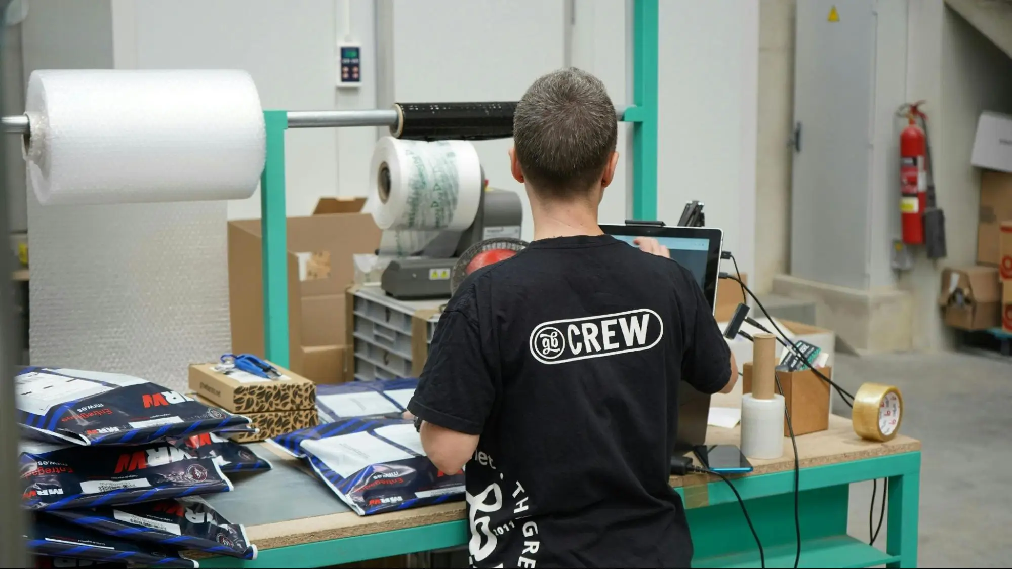 A warehouse worker in a black “CREW” T-shirt stands at a packing station with a laptop, bubble wrap rolls, packing tape, and stacks of blue and white mailer bags.