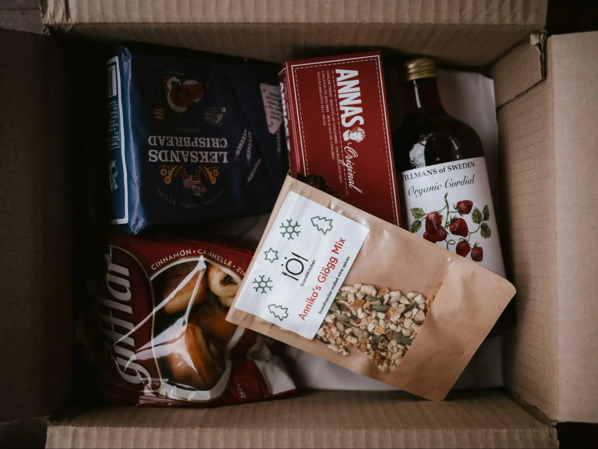 An open cardboard box packed with assorted food products, including a bag of cinnamon biscuits, a box of crispbread, a bottle of organic cordial, and a pouch labeled “Annika’s Glögg Mix.”