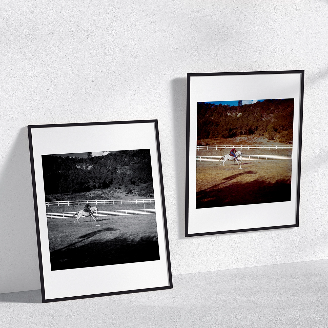 Two framed prints of “Training Grounds” on a light wall: the color version hangs on the right while a black-and-white version leans on the left. Both show a horse and rider mid-jump inside a fenced arena, with trees on the hillside behind.