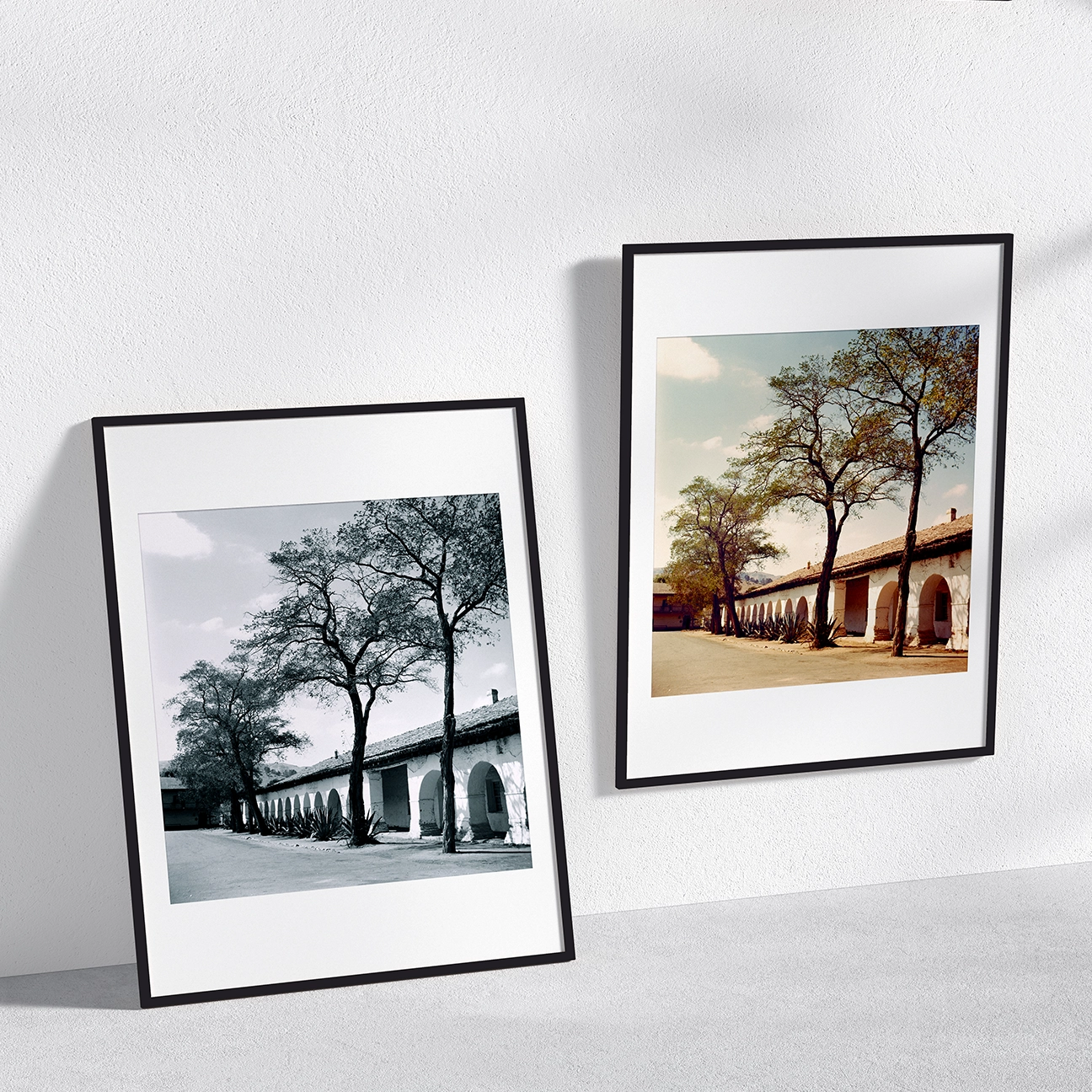 Black-and-white version of “Mission San Juan Bautista,” shown framed on a wall and leaning nearby, featuring a long row of mission arches, stucco walls, and tall trees lining a quiet courtyard.