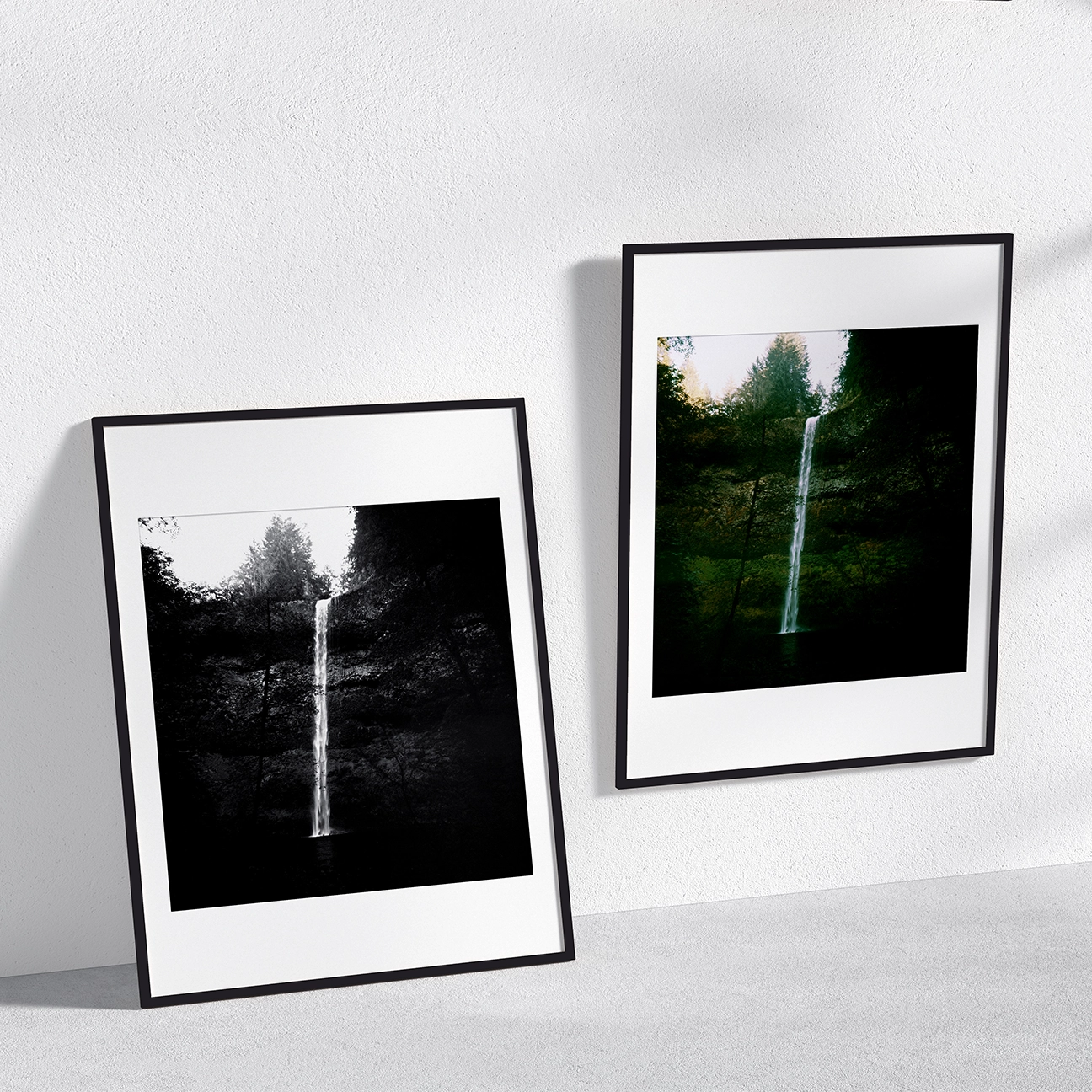 Side-by-side wall mockup of “Silver Creek Falls Waterfall” in black-and-white (left, leaning frame) and color (right, wall-hung). A thin waterfall drops into a dark pool, surrounded by shadowy forest greens and a bright treeline at the top.