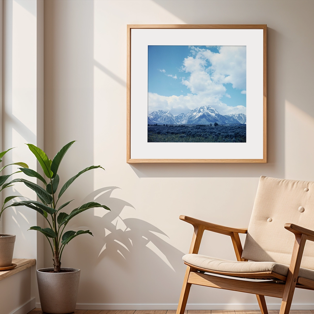 In-room wall mockup featuring a wood-framed print: snowcapped Grand Teton mountains rising above a dark sagebrush plain, with soft, oversized clouds drifting across a clear blue sky.
