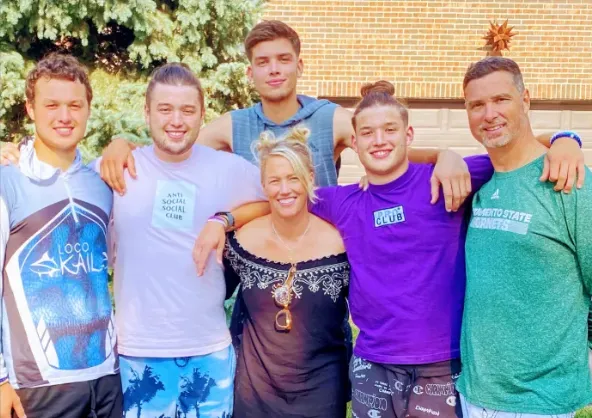 Group of five young men and one woman standing outside smiling, with trees and a brick building in the background.