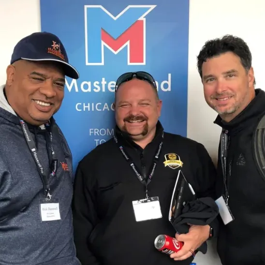 Three men smiling and posing in front of a blue Mastermind Chicago banner, two wearing lanyards and name tags.