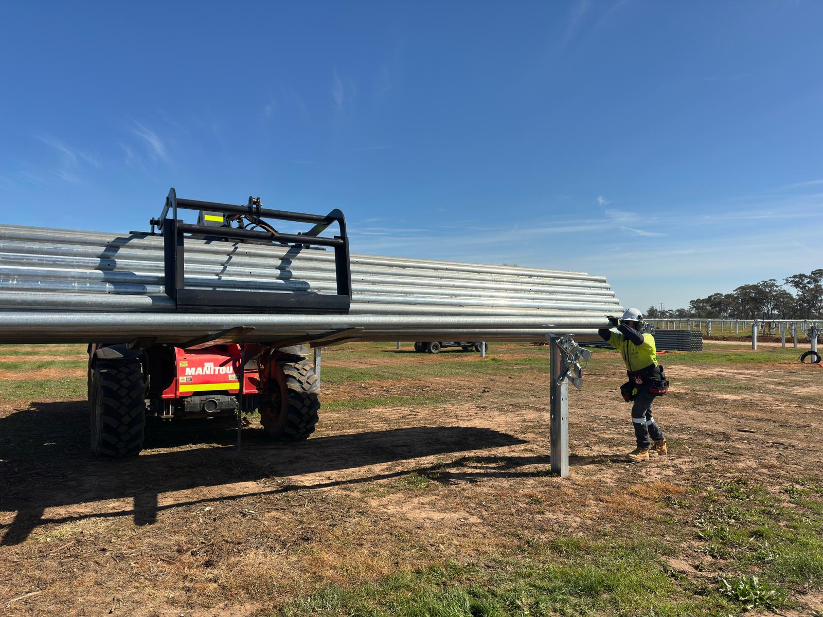 Worker in safety gear handling metal pipes lifted by red Manitou forklift outdoors.