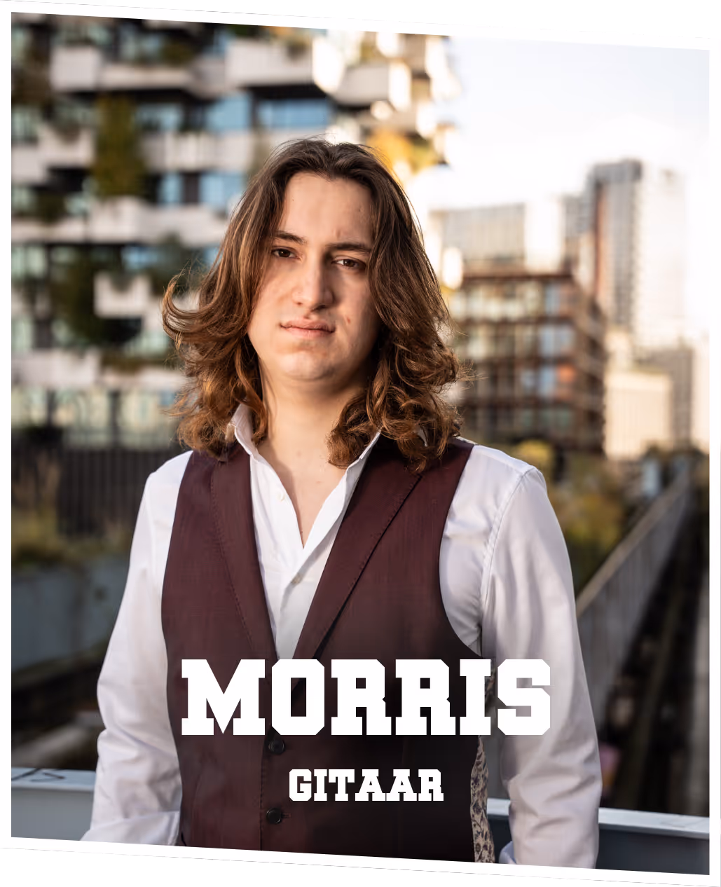 Morris (gitaar) Morris met lang haar en een serieuze blik, in een paars pak met gitaar in de hand, staand voor een moderne gevel.