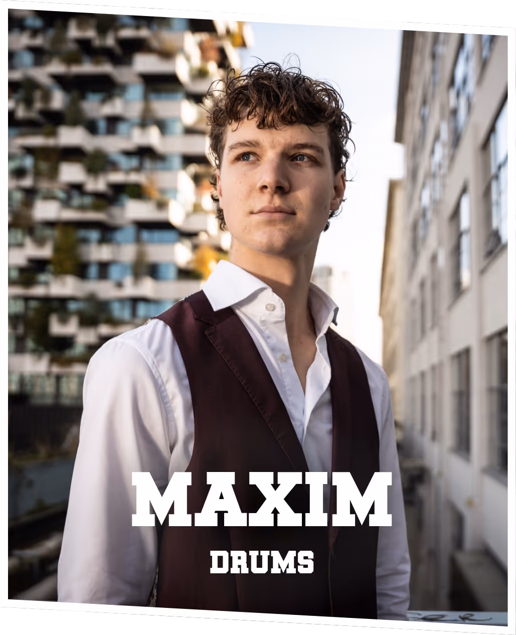 Maxim (drums) Maxim in paars pak, gehurkt bij een auto, terwijl hij zijn drumstokken vast heeft en zijwaarts kijkt.