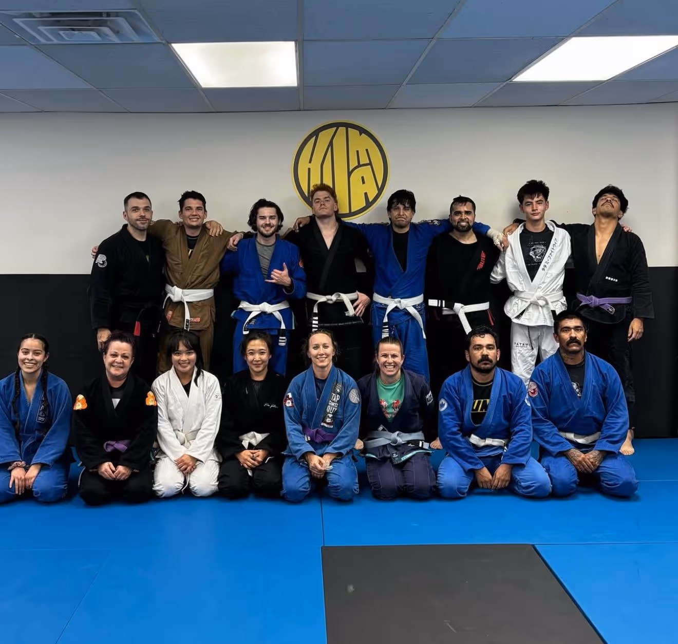 A group of martial artists in varying colored gi uniforms pose for a photo in a training studio with a logo on the wall.