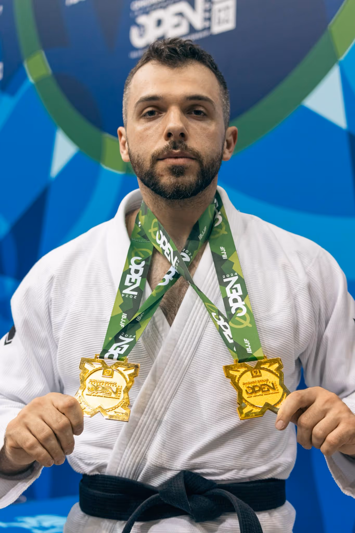 Athlete wearing a judo gi proudly displays two gold medals from the IBJJF Open against a colorful backdrop.