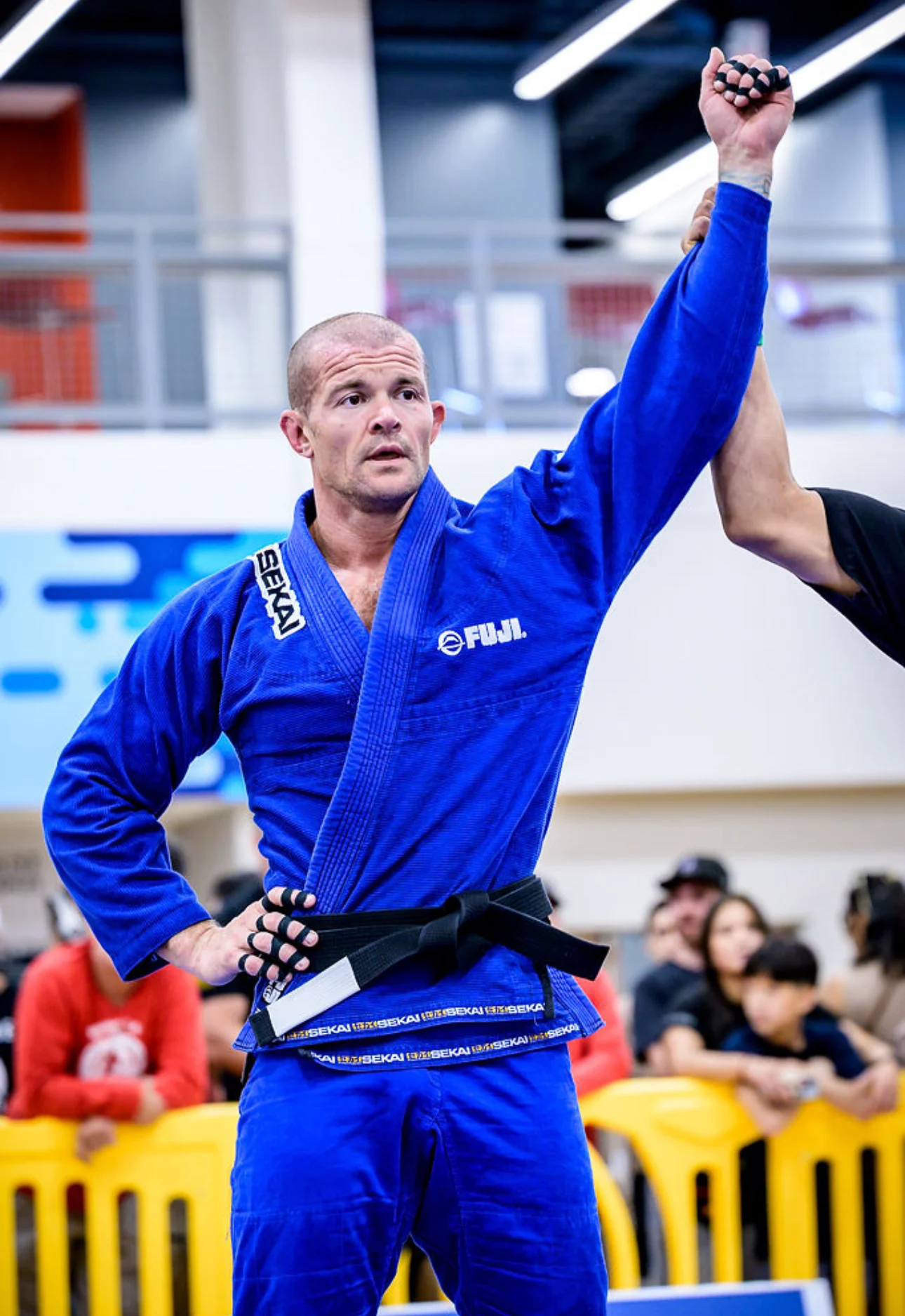 A Brazilian Jiu-Jitsu athlete in a blue gi raises one arm in victory, showcasing determination and focus during a competition.