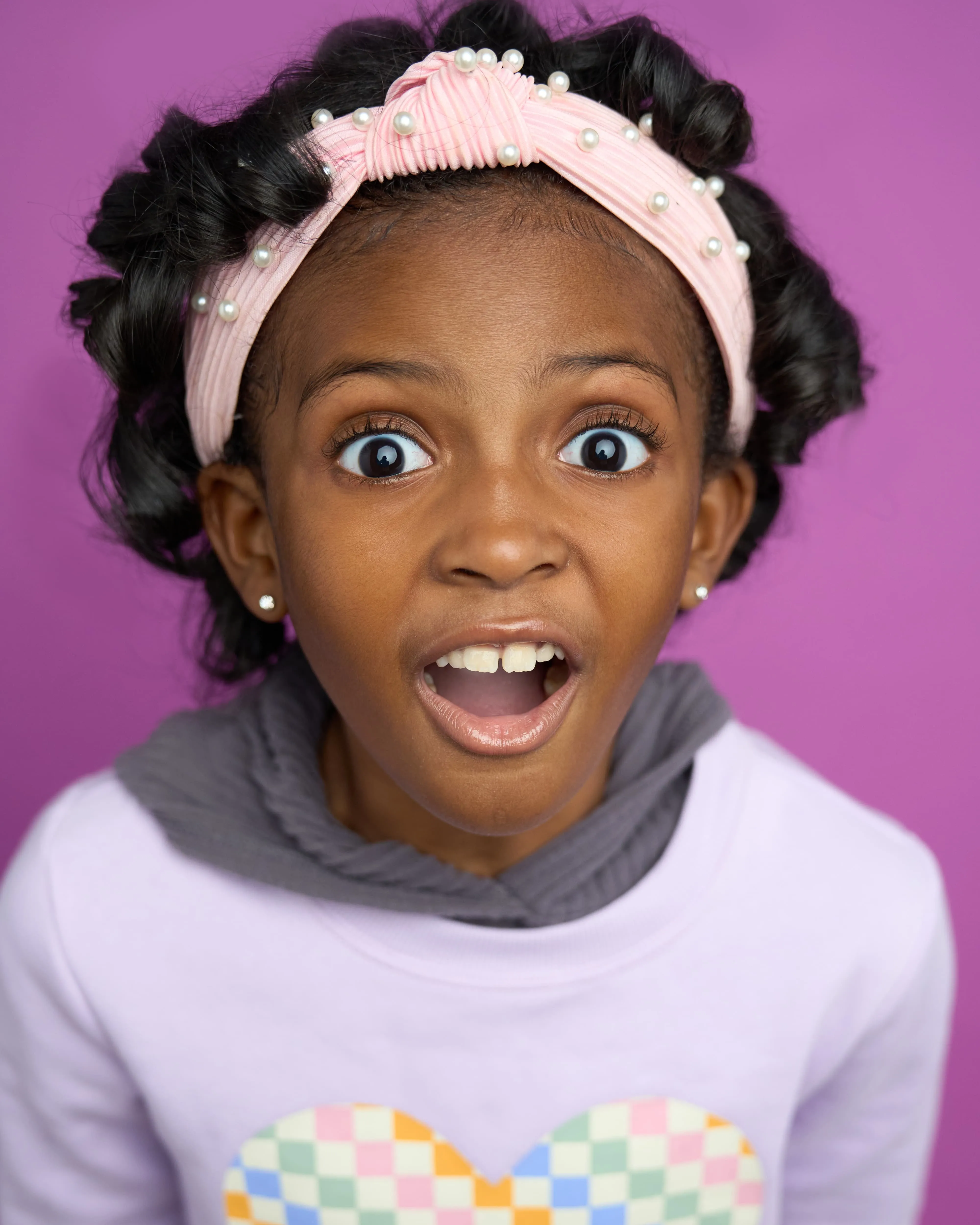 Atlanta kids actor headshot — young Black girl with pearl pink headband, wide-eyed surprised expression in colorful outfit against purple background, expressive commercial kids look.