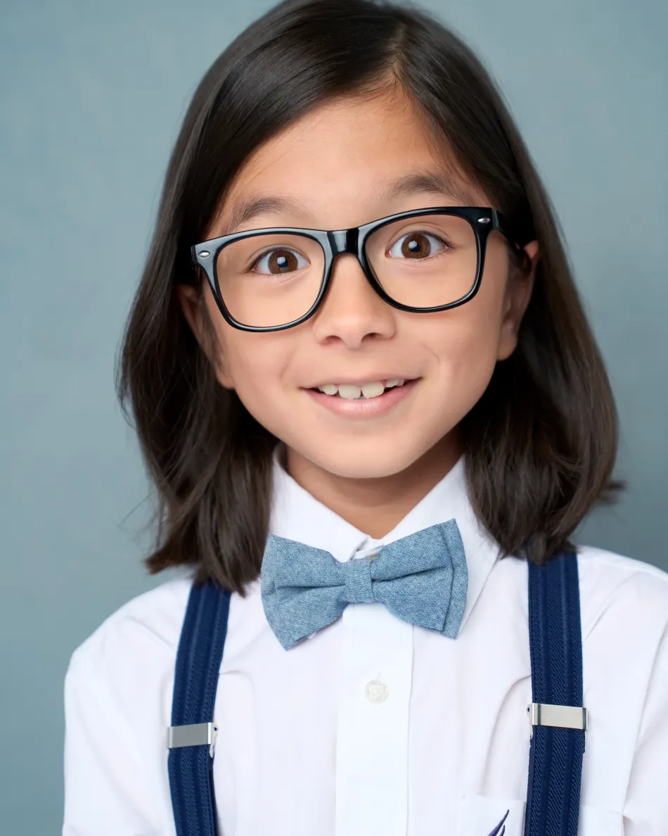 Atlanta kids actor headshot — young boy with glasses, blue bow tie and suspenders, expressive commercial look against light blue background.