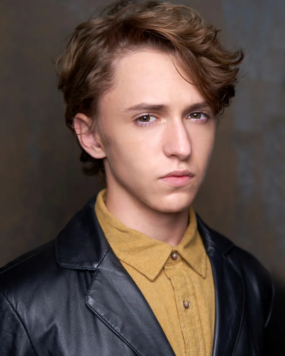 Atlanta theatrical actor headshot — young male with wavy light brown hair, serious dramatic expression in leather jacket and mustard shirt, cinematic studio lighting.