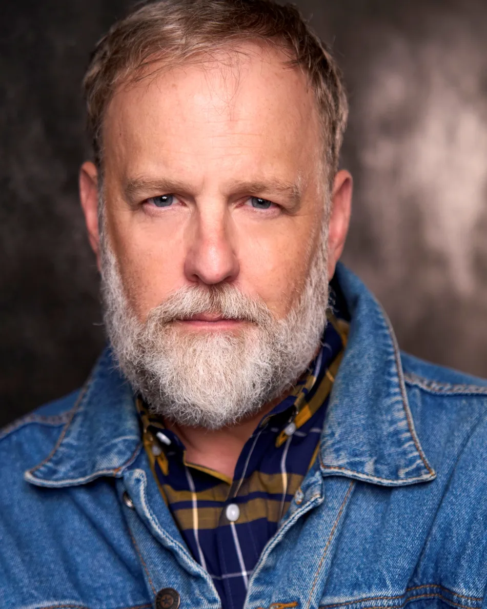 Atlanta theatrical actor headshot — mature male with beard, serious expression, plaid shirt and denim jacket, dramatic studio lighting.