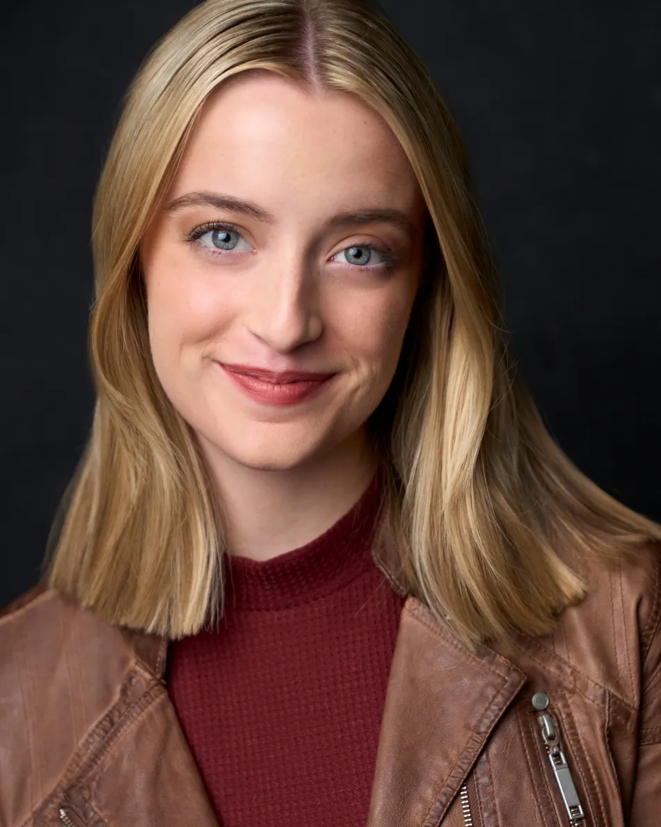 Atlanta theatrical actor headshot — young blonde female theater actress with subtle confident smile, leather jacket and burgundy top against dark studio background.