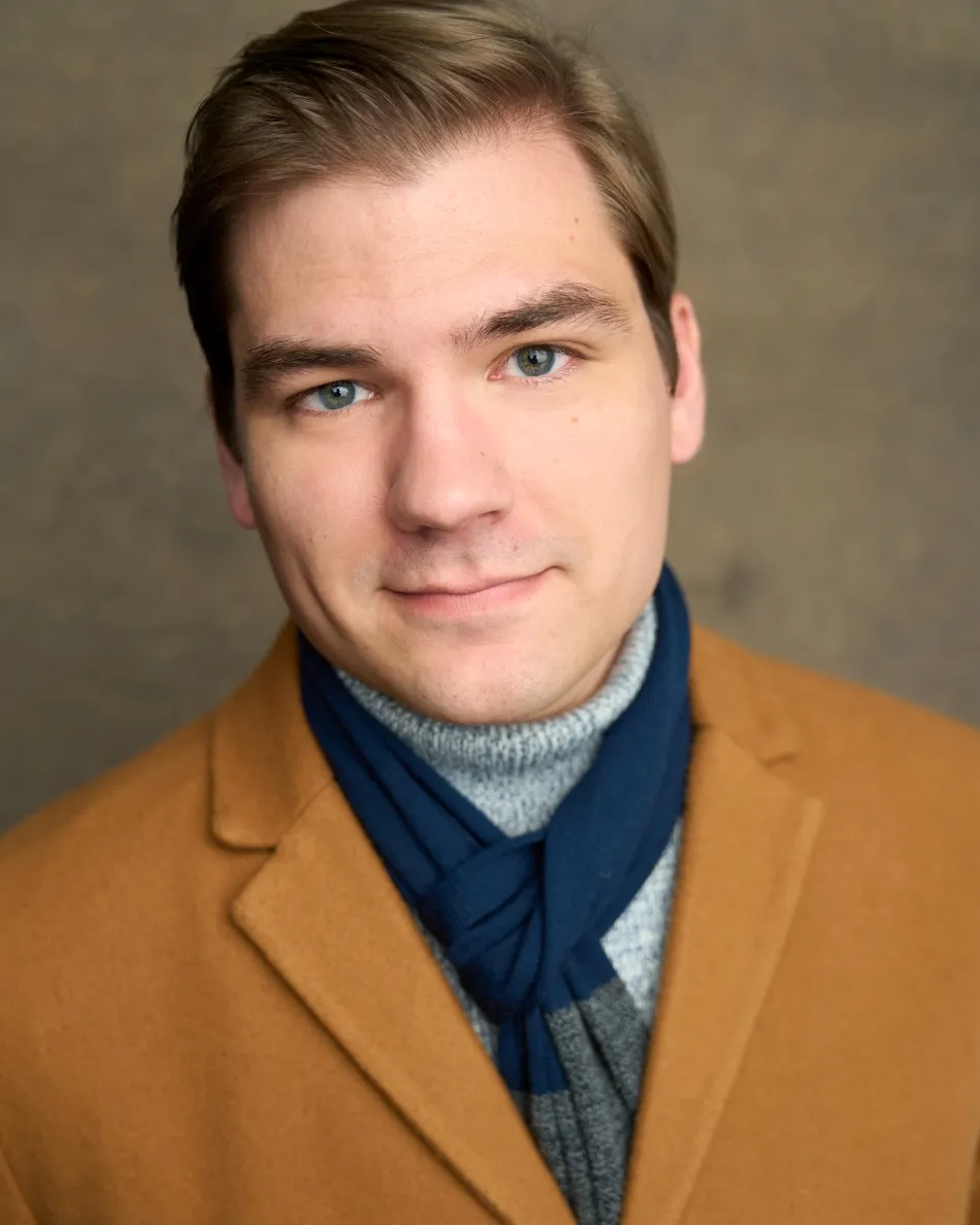Atlanta theatrical actor headshot — young male actor with brown hair, subtle confident smile in tan coat, blue scarf and turtleneck, versatile look for Hallmark, period dramas, or professional roles.