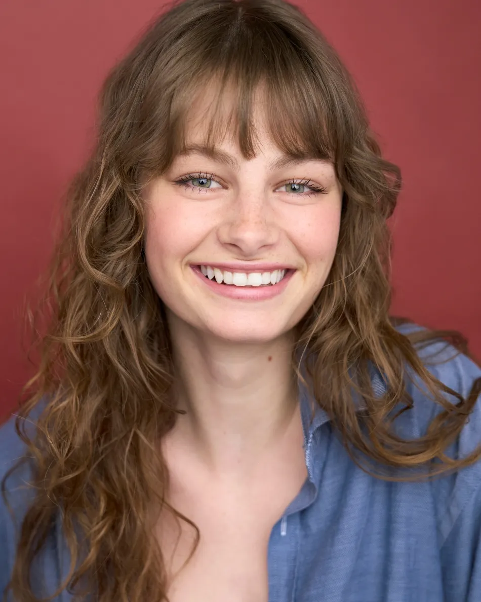 Atlanta musical theater headshot — young female actor with long wavy hair, bright toothy smile in blue shirt against red background, upbeat and expressive theatrical look.
