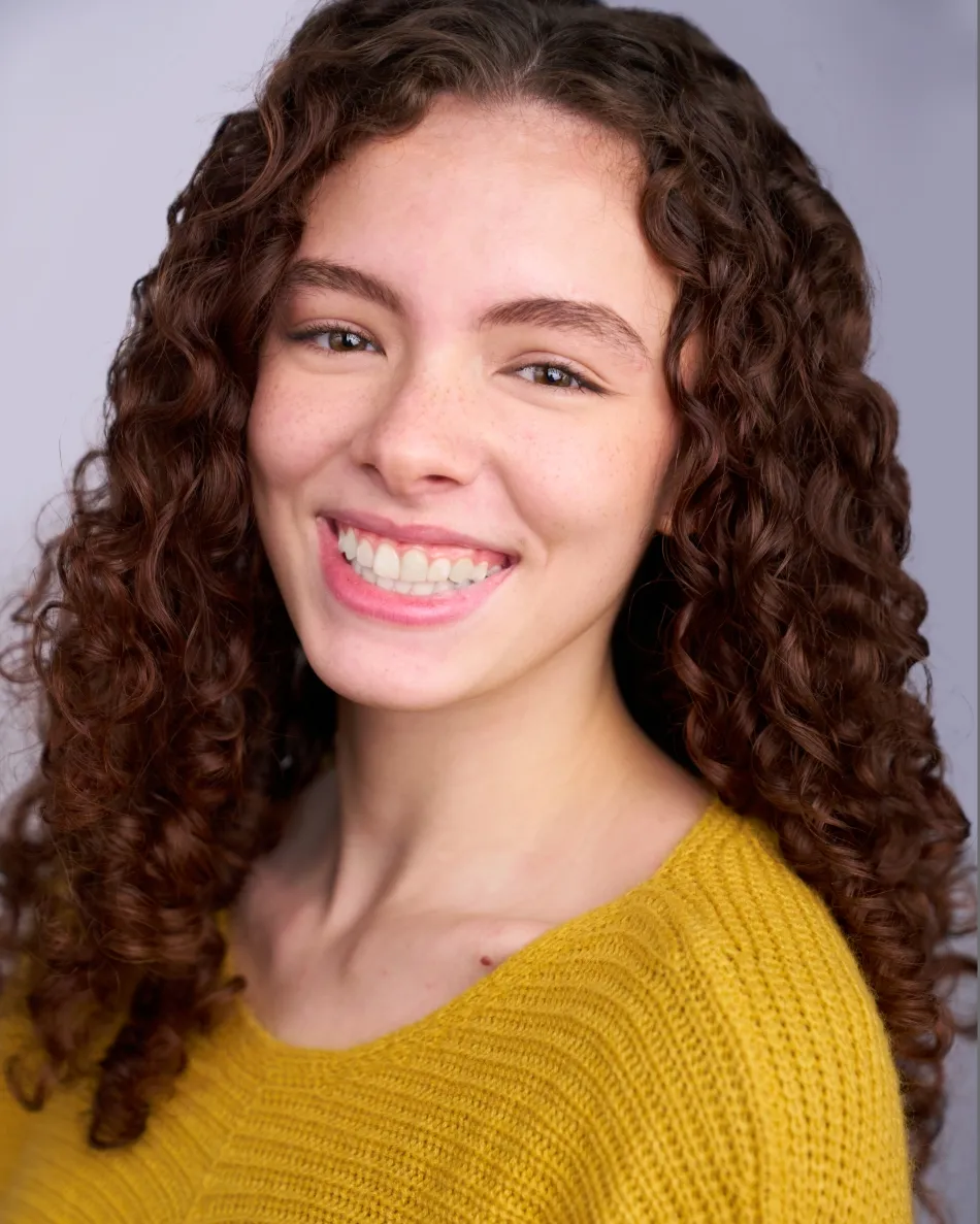 Atlanta musical theater headshot — young female actor with curly hair, bright toothy smile in yellow sweater against light studio background, perfect for musical theater and college auditions.