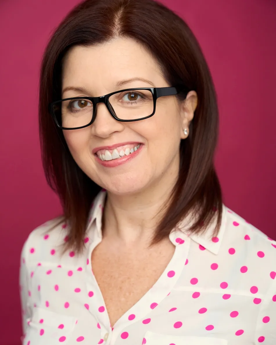 Atlanta commercial actor headshot — middle-aged female with glasses, friendly smile in white blouse with pink polka dots against pink background, approachable and professional look.