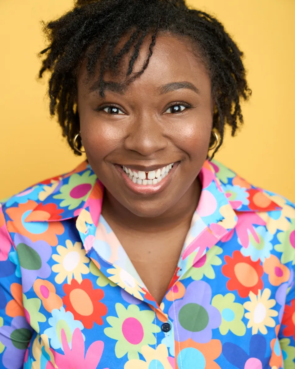 Atlanta commercial actor headshot — female actor with short natural hair, big toothy smile in colorful floral shirt against yellow background, comedic and high-energy look.