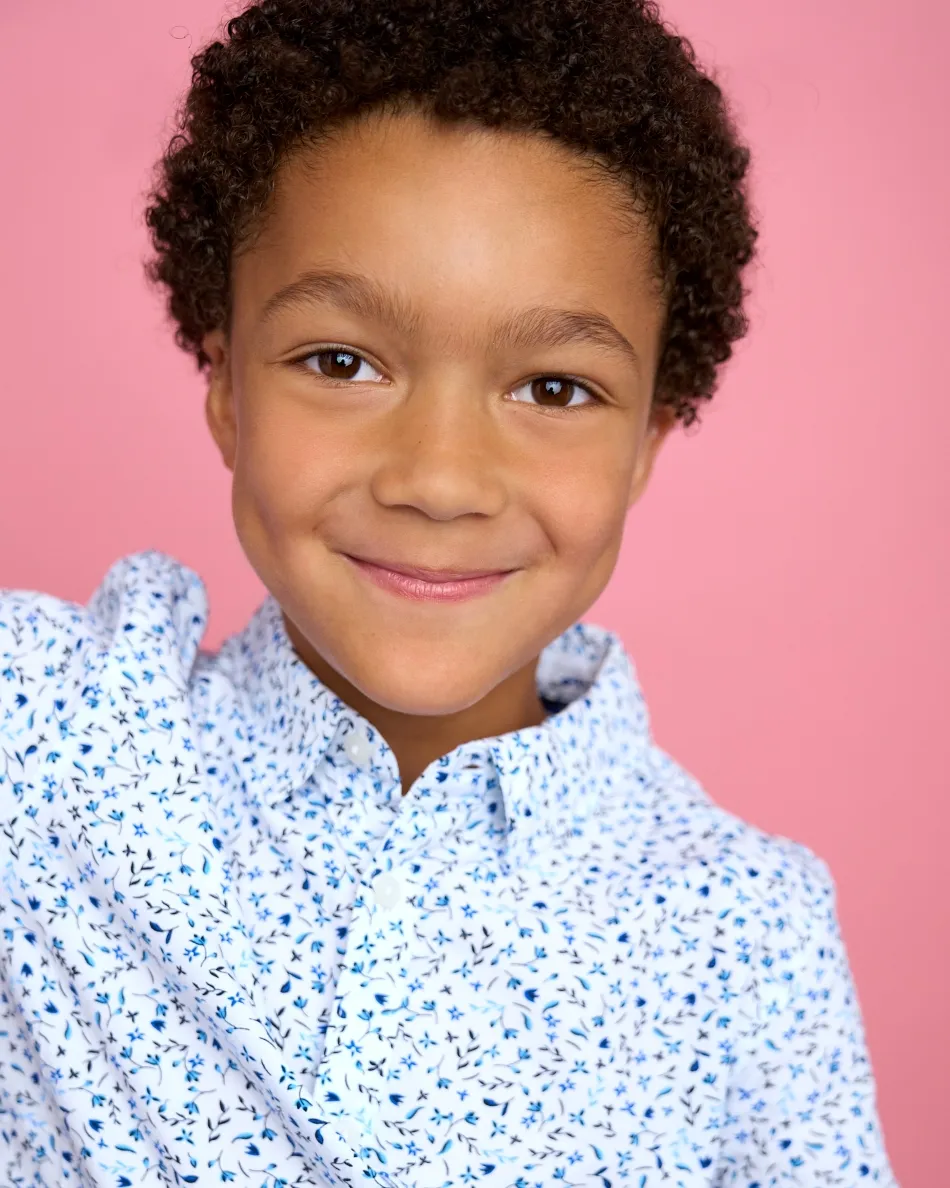 Atlanta kids commercial actor headshot — young boy with curly hair, bright smile in patterned button-up shirt against pink background.