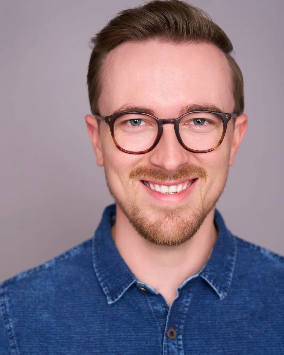 Atlanta college audition headshot — young male actor with glasses and denim shirt, warm toothy smile against neutral background, ideal for musical theater auditions.