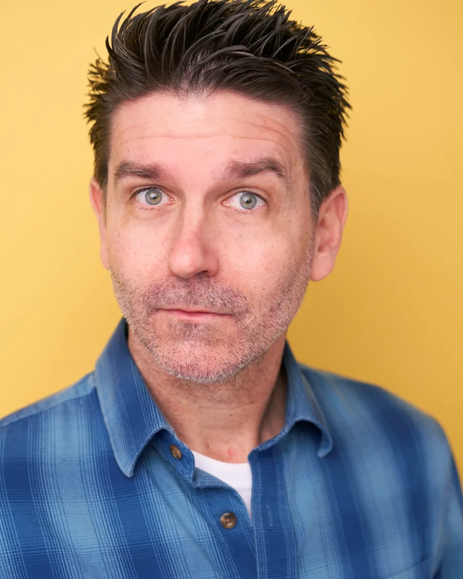 Atlanta comedic commercial actor headshot — male actor with wide-eyed quirky expression, blue plaid shirt against yellow background, character type look.