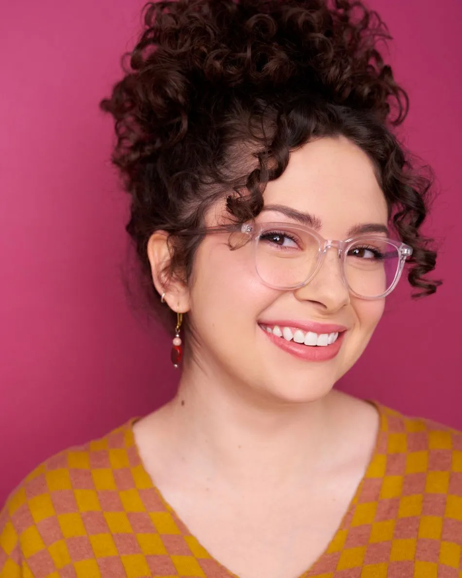 Atlanta commercial actor headshot — young female with curly hair, glasses, bright toothy smile, colorful sweater against pink background.