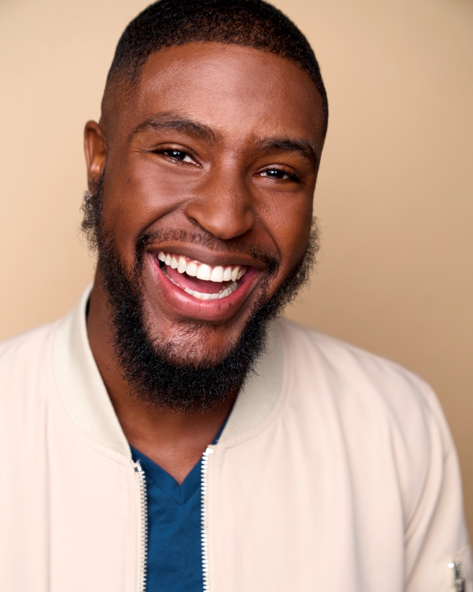 Atlanta commercial actor headshot — young Black male actor with beard, bright toothy smile, casual jacket and blue shirt against beige background.