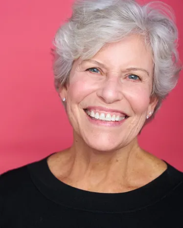 Atlanta commercial actor headshot — mature female actor with short white hair, bright toothy smile in black top against pink background, friendly and welcoming look.