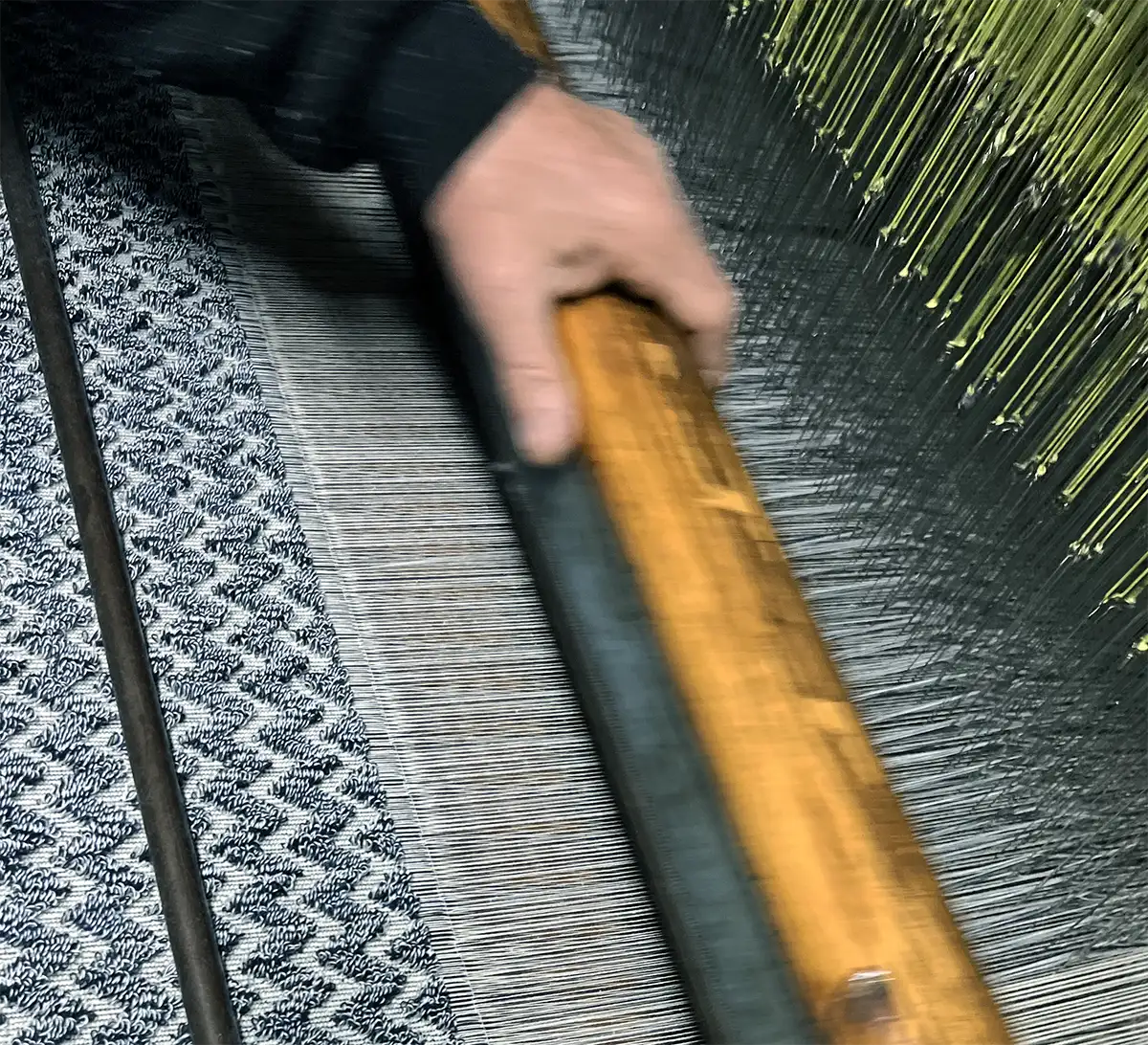 A weaver pulls the beater forward on a traditional shuttle loom, with warp threads visible left to right and weft falling in place in line with the fabric forming