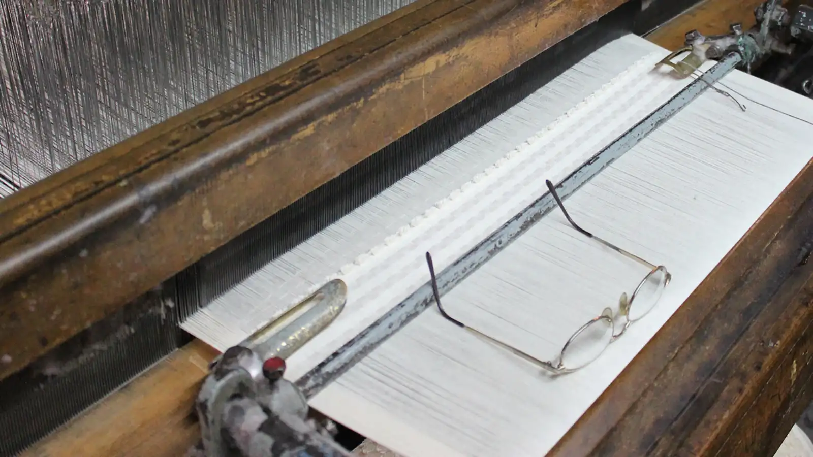 Reading glasses resting on freshly woven fabric beside the reed of a traditional shuttle loom in Türkiye