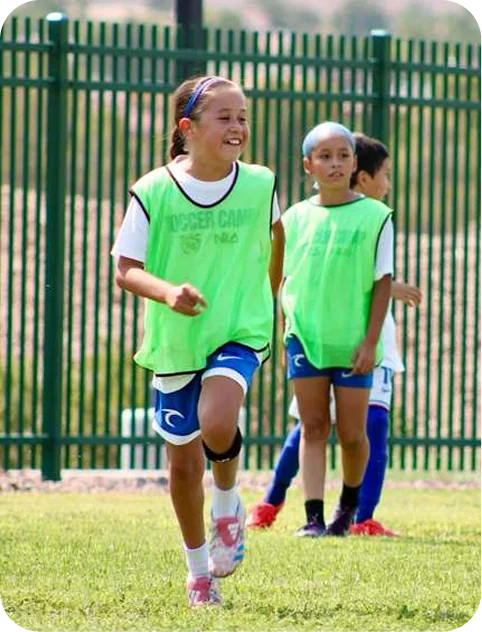 Three children wearing green sports vests playing soccer on a grass field near a green metal fence.